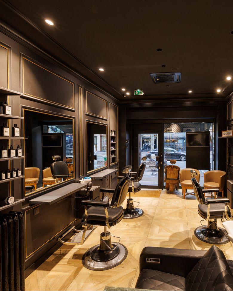 Intérieur élégant d’un salon de coiffure/barbier : plusieurs fauteuils en cuir noir devant de grands miroirs, sol en parquet clair et étagères de produits, avec vue sur la vitrine donnant sur la rue.