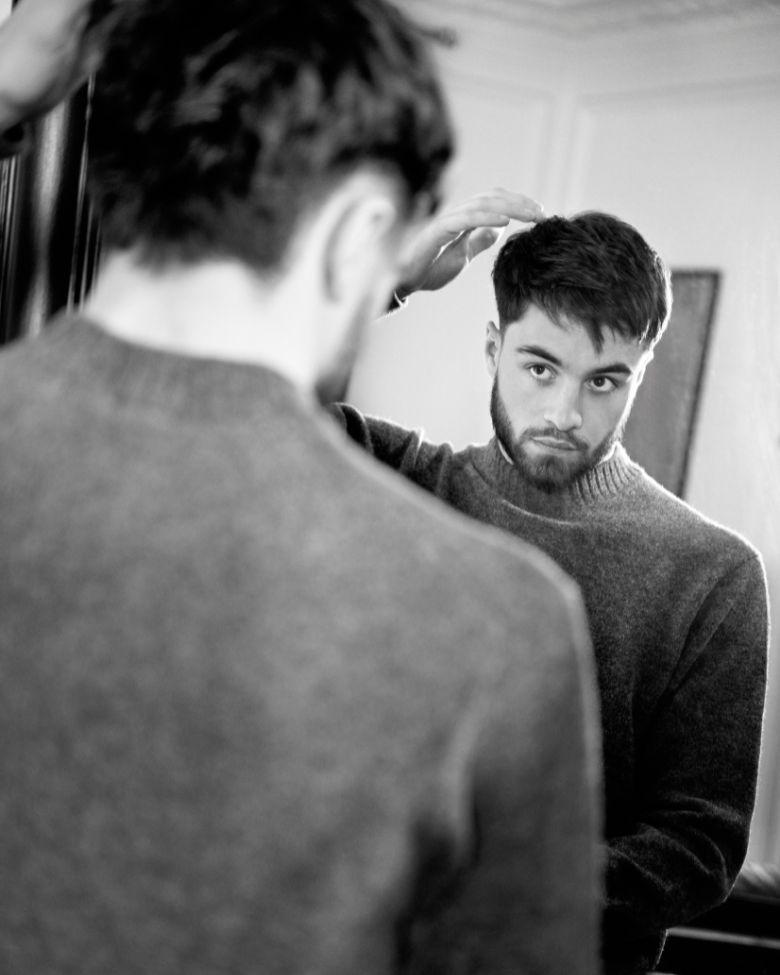 Jules Bouyer, homme se regardant dans un miroir en ajustant ses cheveux, portant un pull en laine gris, photo en noir et blanc.